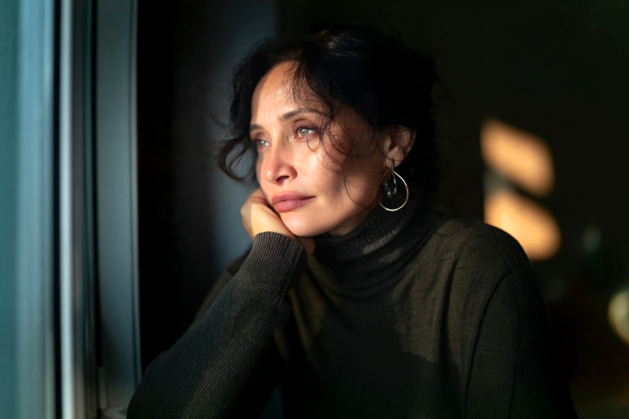 Woman with dark hair and hoop earrings gazes thoughtfully out a window, resting her chin on her hand. She wears a dark turtleneck, and soft sunlight falls across her face. The mood appears pensive and reflective.