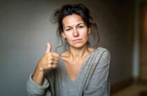 A woman with messy hair and a neutral expression gives a thumbs-up gesture. She is wearing a loose gray sweater and is positioned against a plain background.