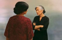 Two women stand facing each other. One, with gray hair in a bun and arms crossed, looks stern. The other, seen from behind, wears a red patterned dress. The background is soft and muted.