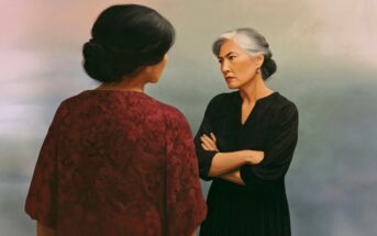 Two women stand facing each other. One, with gray hair in a bun and arms crossed, looks stern. The other, seen from behind, wears a red patterned dress. The background is soft and muted.