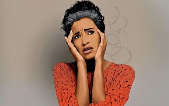 A woman in a red patterned shirt holds her face with both hands and looks worried or anxious, with a neutral background behind her.