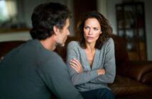 A woman with crossed arms looks seriously at a man seated across from her. They appear to be having a tense conversation in a dimly lit living room.