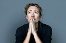A person with light hair pulled back looks upwards with hands pressed together in front of their face, appearing thoughtful or worried, against a plain gray background.
