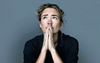 A person with light hair pulled back looks upwards with hands pressed together in front of their face, appearing thoughtful or worried, against a plain gray background.