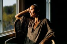 A person sits in a wooden chair by a sunlit window, resting their arm on the back, and gazing outside with a gentle smile. Warm sunlight highlights their face and loose, earth-toned clothing.