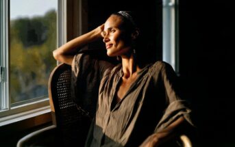 A person sits in a wooden chair by a sunlit window, resting their arm on the back, and gazing outside with a gentle smile. Warm sunlight highlights their face and loose, earth-toned clothing.