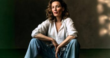 A woman with wavy hair sits on the floor in a relaxed pose, wearing a white button-up shirt and blue jeans, with a neutral, softly lit background.