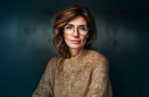 A woman with wavy, reddish-brown hair and glasses wears a tan, chunky knit sweater. She sits against a dark, blurred background, looking calmly at the camera with a slight, confident smile.
