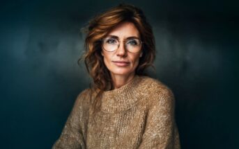 A woman with wavy, reddish-brown hair and glasses wears a tan, chunky knit sweater. She sits against a dark, blurred background, looking calmly at the camera with a slight, confident smile.