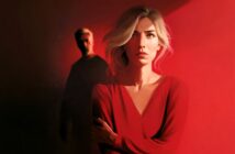 A woman in red stands in dramatic lighting, looking worried. A man in dark clothes is blurred in the background, partially in shadow.
