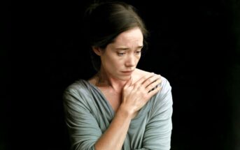 A woman in a gray top stands against a dark background, looking down with a sad expression. One hand is pressed to her chest, and her hair is tied back.