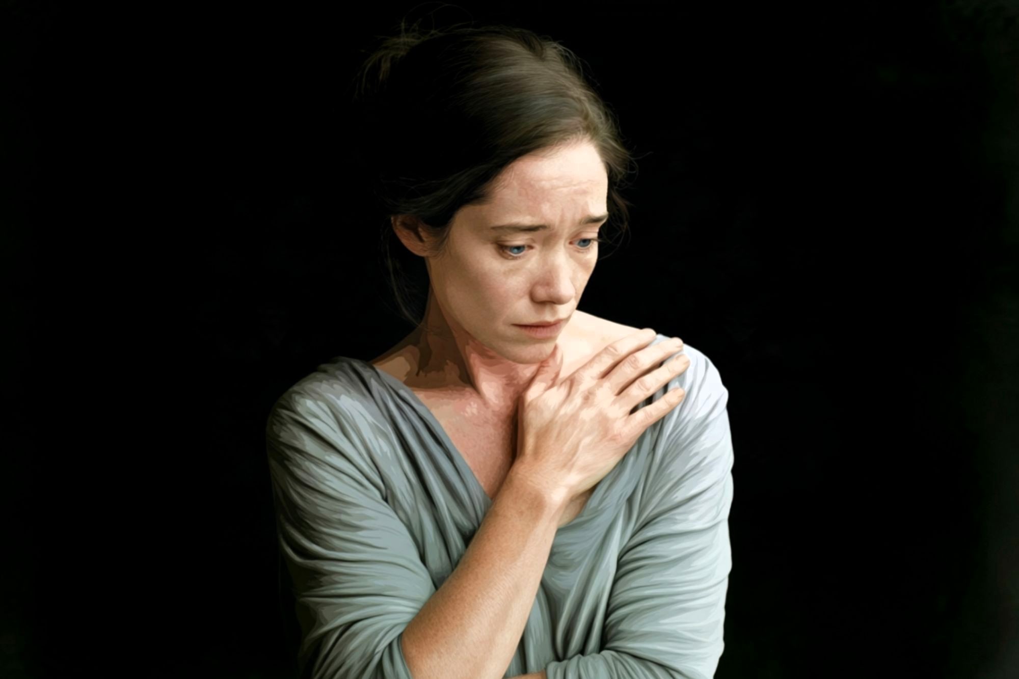 A woman in a gray top stands against a dark background, looking down with a sad expression. One hand is pressed to her chest, and her hair is tied back.
