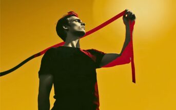 A person in a black shirt holds a red measuring tape confidently against a bright yellow background, looking upward toward the light.