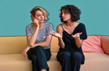 Two women sit on a beige couch against a teal wall. One, in a striped shirt, looks thoughtful with her hand on her face. The other, in a black shirt, gestures with her hands while speaking.