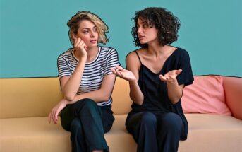 Two women sit on a beige couch against a teal wall. One, in a striped shirt, looks thoughtful with her hand on her face. The other, in a black shirt, gestures with her hands while speaking.