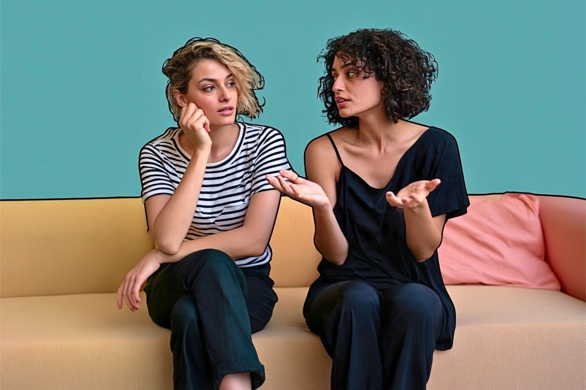 Two women sit on a beige couch against a teal wall. One, in a striped shirt, looks thoughtful with her hand on her face. The other, in a black shirt, gestures with her hands while speaking.