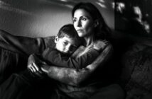 A woman sits on a couch, holding a young boy in her arms. Both have serious expressions and stare into the distance. The image is in black and white, with soft light and shadows on the wall behind them.