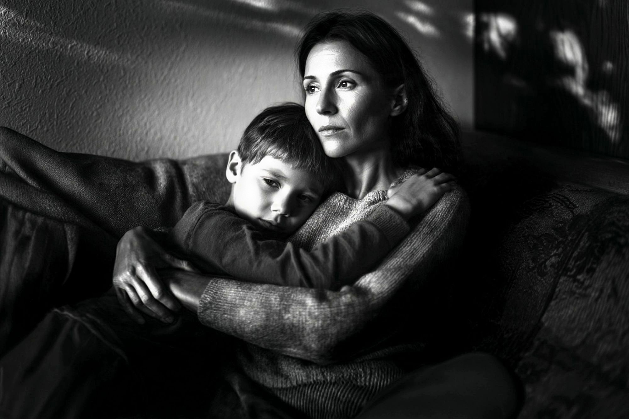A woman sits on a couch, holding a young boy in her arms. Both have serious expressions and stare into the distance. The image is in black and white, with soft light and shadows on the wall behind them.