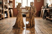 A wooden sculpture of a woman and man holding hands, each with a puzzle piece cutout that fits together, stands on a wooden floor in a sunlit, cozy workshop.