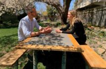 A man and a woman sit across from each other at a wooden picnic table in a sunlit garden.