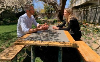 A man and a woman sit across from each other at a wooden picnic table in a sunlit garden.