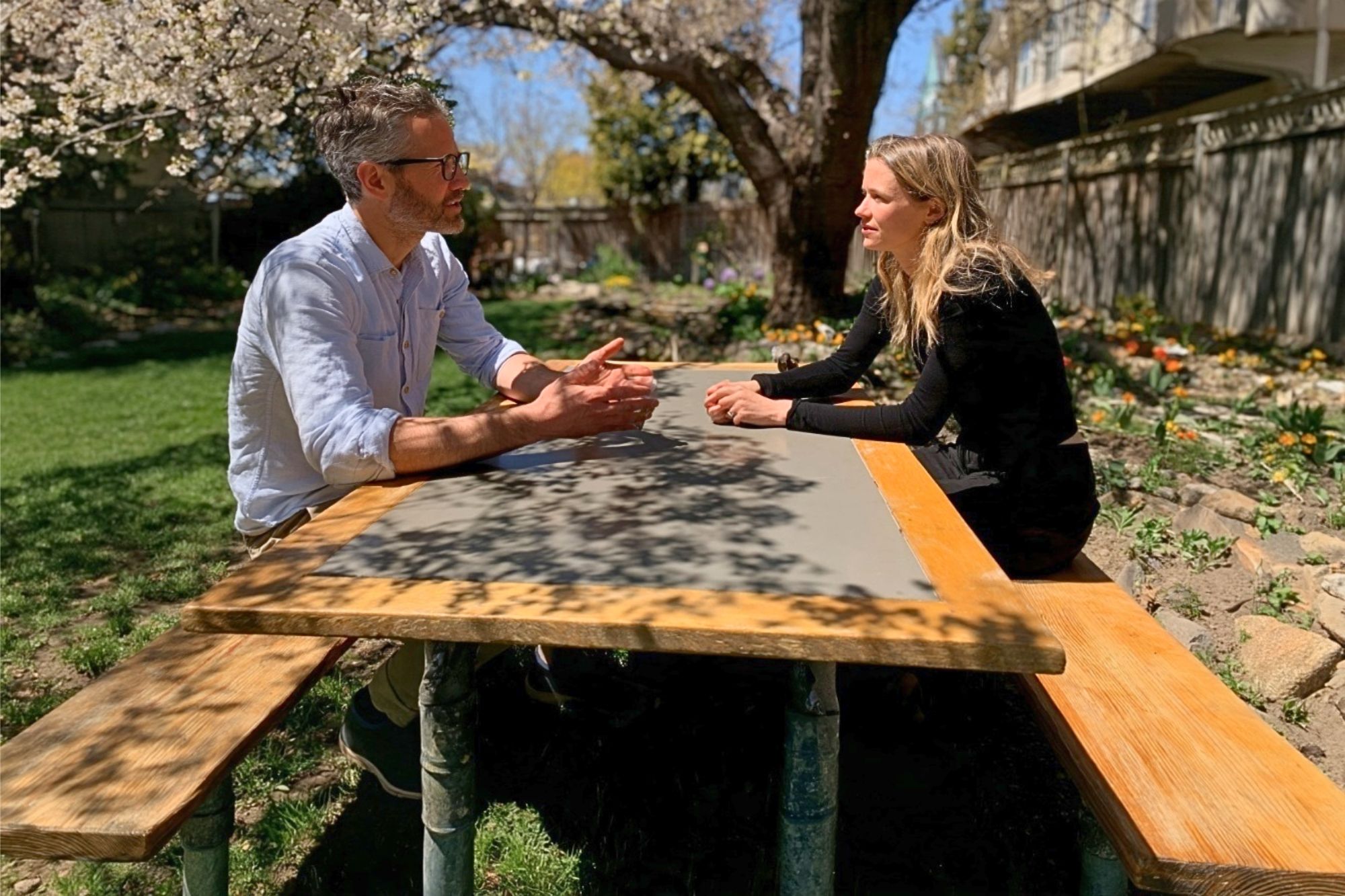 A man and a woman sit across from each other at a wooden picnic table in a sunlit garden.
