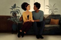 A couple sits together on a sofa, holding hands and looking at each other.