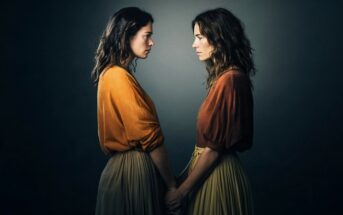Two women with similar features, wearing orange tops and beige skirts, stand face-to-face and hold hands against a dark gradient background.