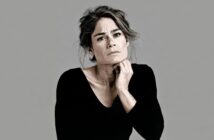 A portrait of a woman in a black long-sleeved shirt with her hand resting under her chin, looking directly at the camera.
