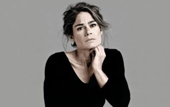 A portrait of a woman in a black long-sleeved shirt with her hand resting under her chin, looking directly at the camera.