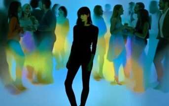 A woman stands alone in the center of a social gathering, surrounded by groups of people talking, with colorful blurred lights creating a dreamy, isolated effect.