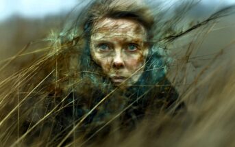 A woman with blue eyes stares ahead, surrounded by tall dry grass blowing in the wind. Her face is partly obscured by the grass, creating a dramatic and moody outdoors scene.