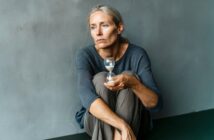A woman with gray hair sits on the floor holding a small hourglass and looking away with a pensive expression.