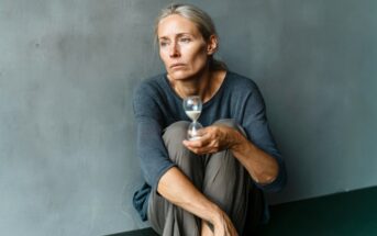 A woman with gray hair sits on the floor holding a small hourglass and looking away with a pensive expression.