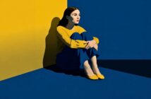 A woman in a yellow top and blue pants sits on the floor in the corner of a room with yellow and blue walls.