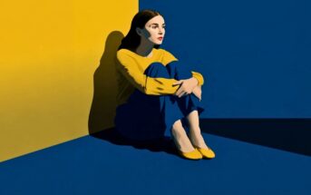 A woman in a yellow top and blue pants sits on the floor in the corner of a room with yellow and blue walls.