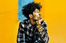 A young man with curly hair wearing a plaid shirt rests his chin on his hand while looking off to the side against a yellow and blue background.