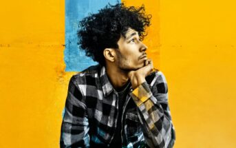 A young man with curly hair wearing a plaid shirt rests his chin on his hand while looking off to the side against a yellow and blue background.