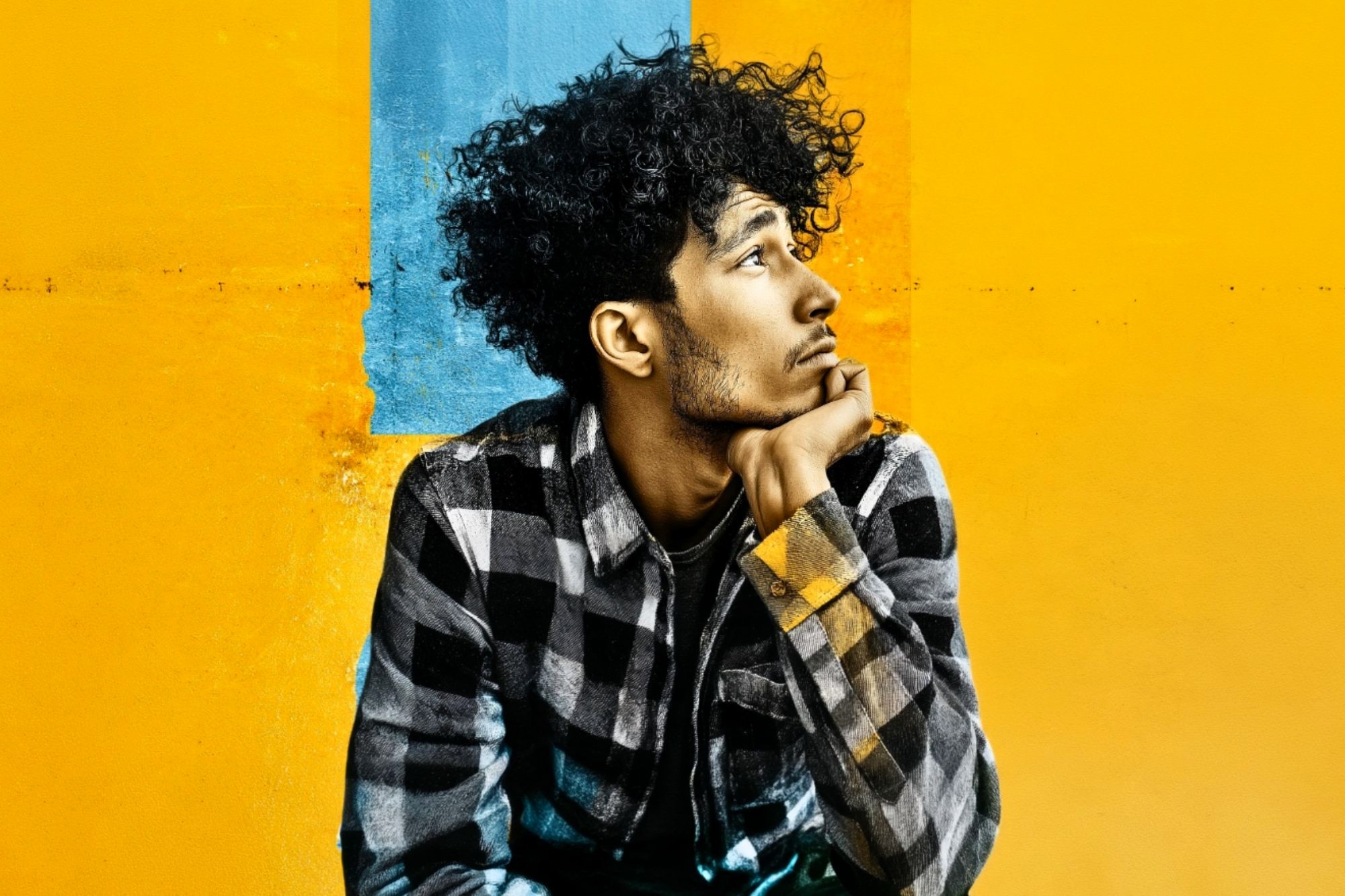 A young man with curly hair wearing a plaid shirt rests his chin on his hand while looking off to the side against a yellow and blue background.