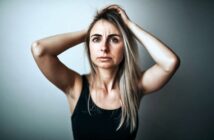 A woman in a black tank top with her hands raised to her head, looking forward with a concerned expression.