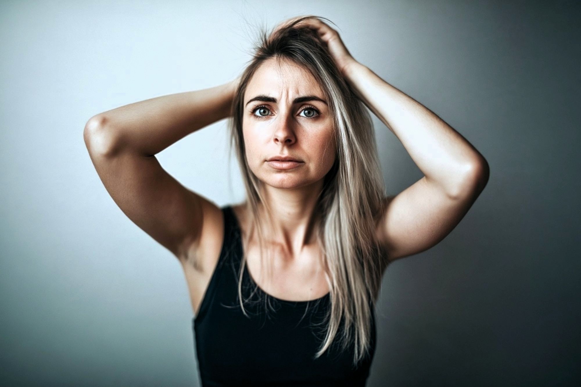 A woman in a black tank top with her hands raised to her head, looking forward with a concerned expression.