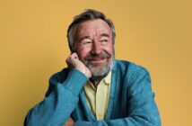 A smiling older man with a grey beard rests his chin on his hand, looking off to the side against a yellow background.