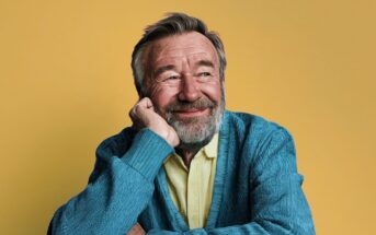 A smiling older man with a grey beard rests his chin on his hand, looking off to the side against a yellow background.