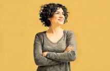 A smiling woman with curly hair stands with her arms crossed against a yellow background.
