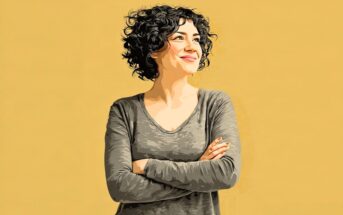A smiling woman with curly hair stands with her arms crossed against a yellow background.