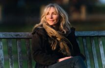 A woman in a dark jacket and scarf sitting on a wooden bench, looking off into the distance.