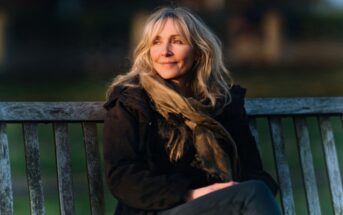 A woman in a dark jacket and scarf sitting on a wooden bench, looking off into the distance.