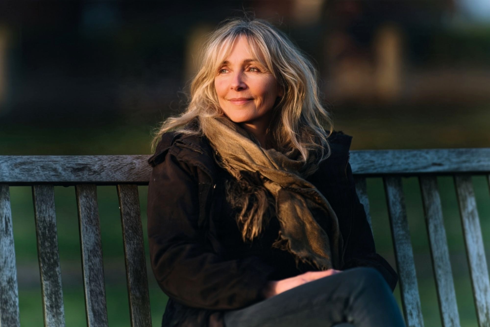 A woman in a dark jacket and scarf sitting on a wooden bench, looking off into the distance.