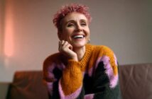 A smiling person with short, pink curly hair wears a chunky knit sweater with large blocks of orange, pink, and dark grey.