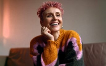 A smiling person with short, pink curly hair wears a chunky knit sweater with large blocks of orange, pink, and dark grey.